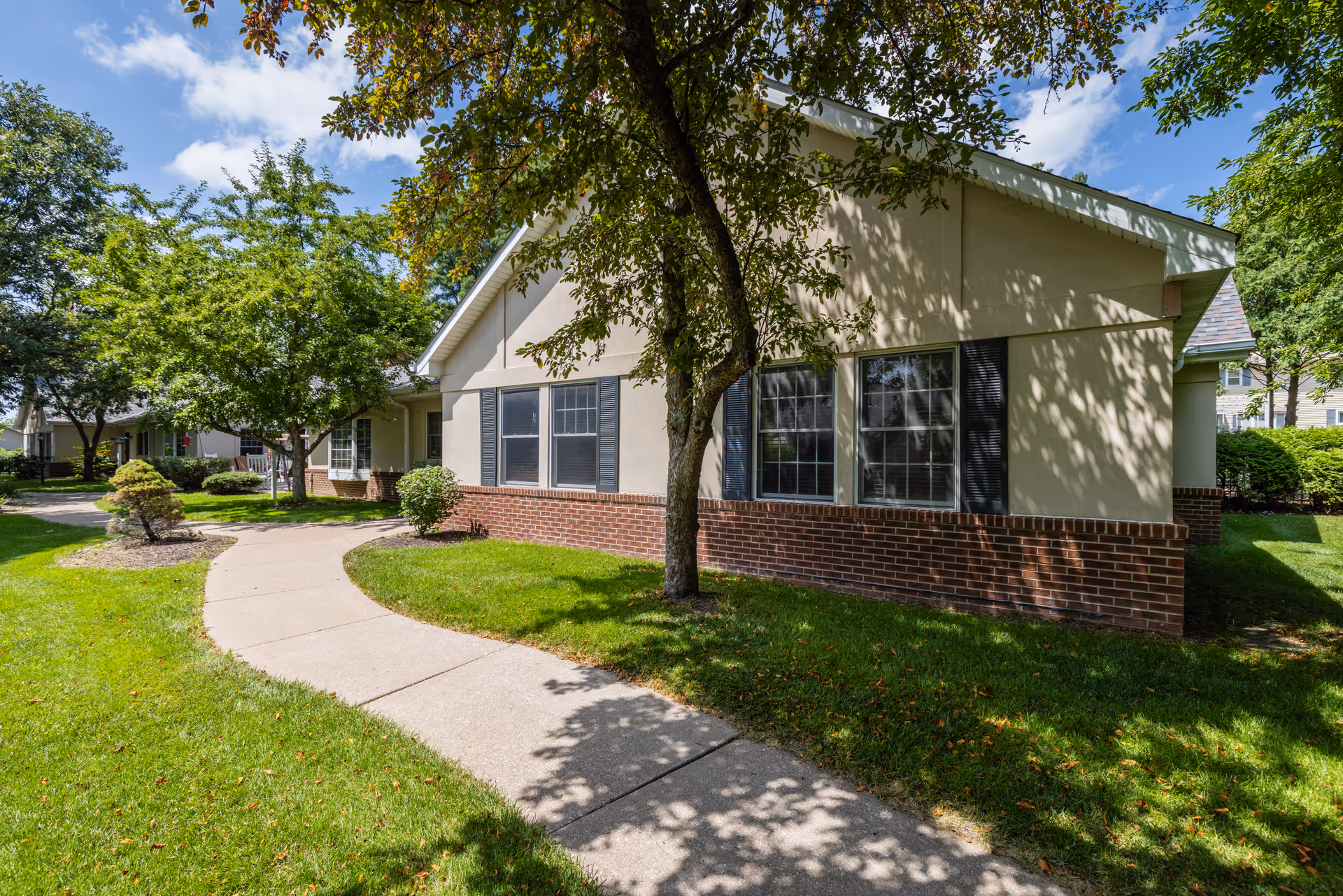 Exterior front of a single-story memory care building with a curved sidewalk, lawn, trees, and brick foundation.