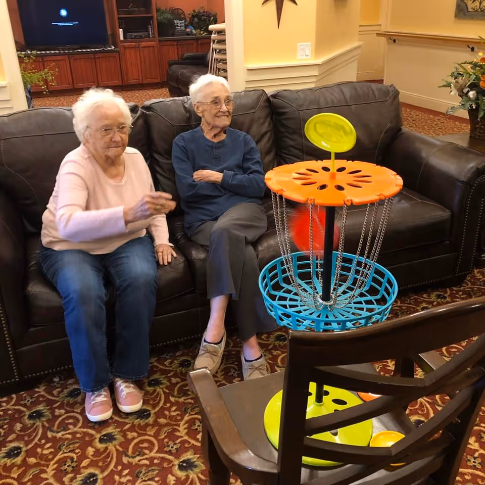 Two elderly women sit on a leather couch in a communal living room playing a small tabletop disc-golf style game.