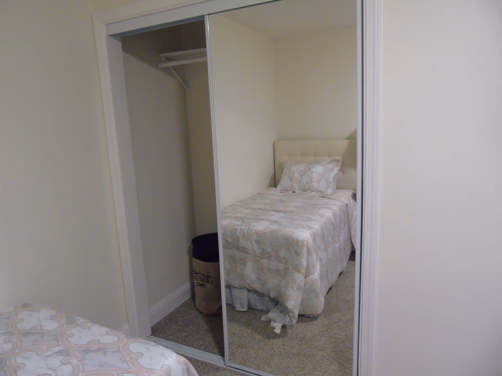 A small bedroom with a single bed covered in a patterned bedspread and matching pillow. There is a mirrored sliding closet door reflecting the bed and a laundry basket inside the closet. The walls are painted light beige and the floor is carpeted.