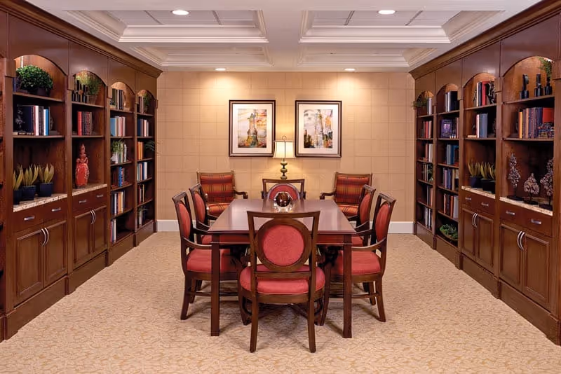A cozy library/meeting room with a wooden table surrounded by red-upholstered chairs and built-in bookshelves lining both walls.
