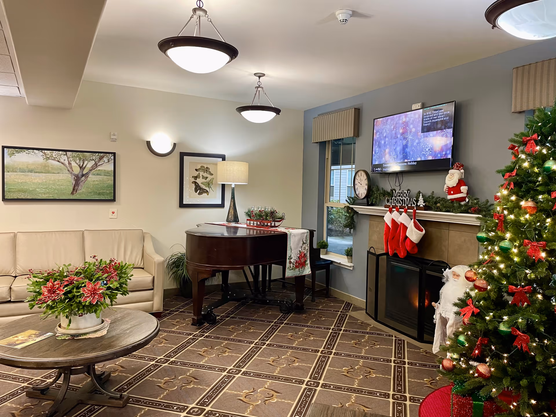 A cozy living room decorated for Christmas with a decorated Christmas tree, stockings hanging on a fireplace mantel, a flat-screen TV mounted above the fireplace, a grand piano with a lamp on it, a beige sofa, a round wooden coffee table with a festive floral arrangement, and framed artwork on the walls.