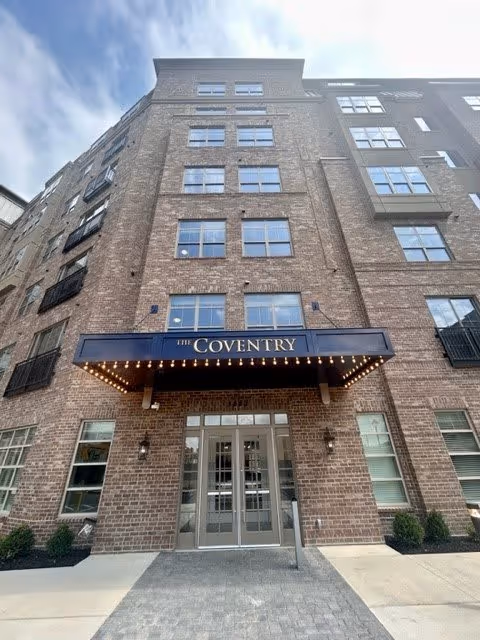 Exterior view of a multi-story brick building with a canopy above the entrance that reads 'The Coventry'. The building has multiple windows and small shrubs near the entrance.