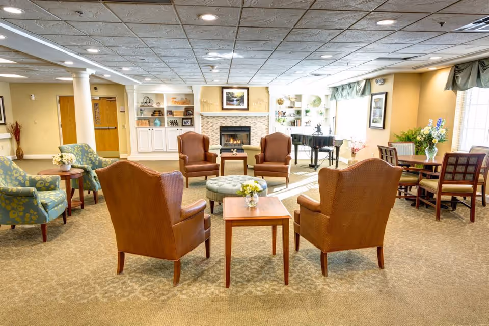 A bright and spacious senior living facility common area with comfortable seating including brown armchairs and patterned blue chairs arranged around small wooden tables. A fireplace with a framed picture above it is centered on the far wall, flanked by built-in white shelves displaying decorative items. To the right, there is a black grand piano near large windows with green valances, and a dining table with chairs and a vase of flowers. The room has a beige carpet and a white tiled ceiling with recessed lighting.