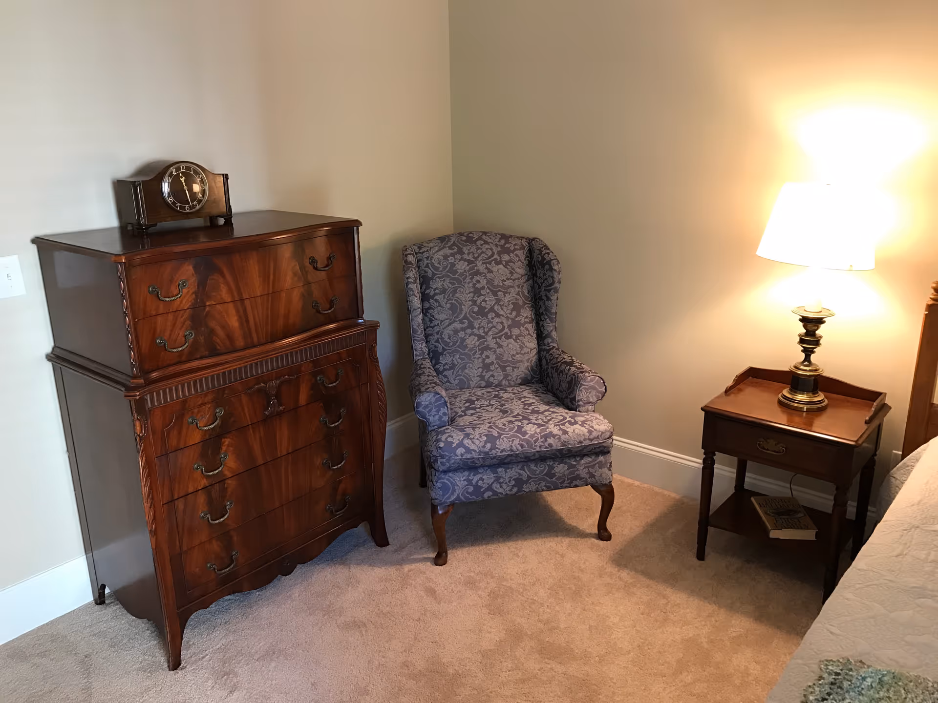 A cozy corner of a bedroom featuring a vintage wooden chest of drawers with a classic clock on top, a patterned upholstered armchair, and a wooden nightstand with a brass lamp emitting warm light. The floor is carpeted and the walls are painted beige.