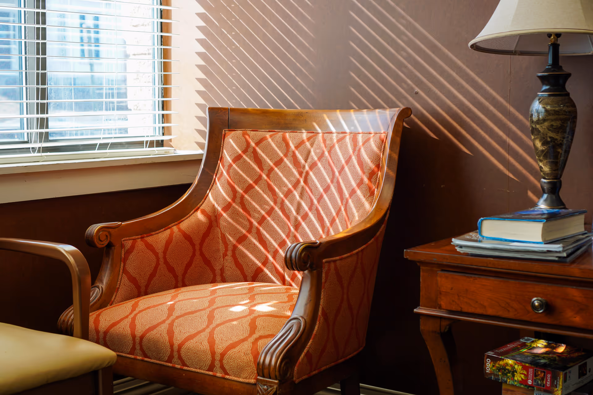 A cozy corner with an upholstered armchair featuring a red and beige patterned fabric, positioned next to a wooden side table with a lamp, books, and a puzzle box underneath. Sunlight streams through window blinds, casting striped shadows on the chair and wall.