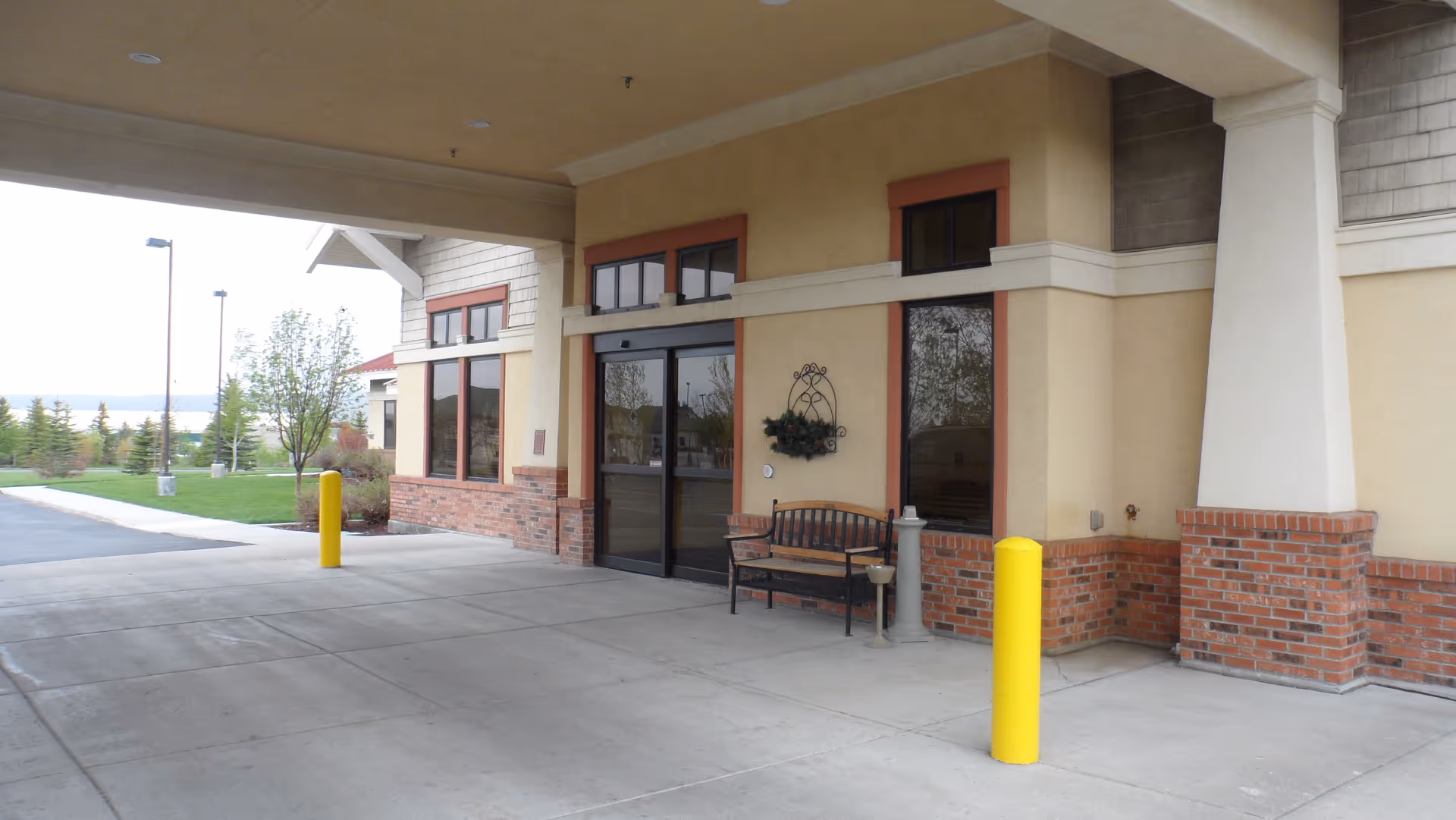 Covered entrance area of a building with double glass doors, a bench, and yellow bollards. The building exterior features beige walls with brick accents and large windows. There is a grassy area with trees and lamp posts visible in the background.