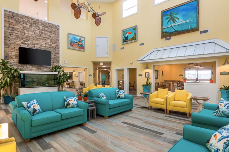 Bright communal living room with teal sofas, yellow armchairs, coastal artwork, a TV above a stone fireplace, and large windows.
