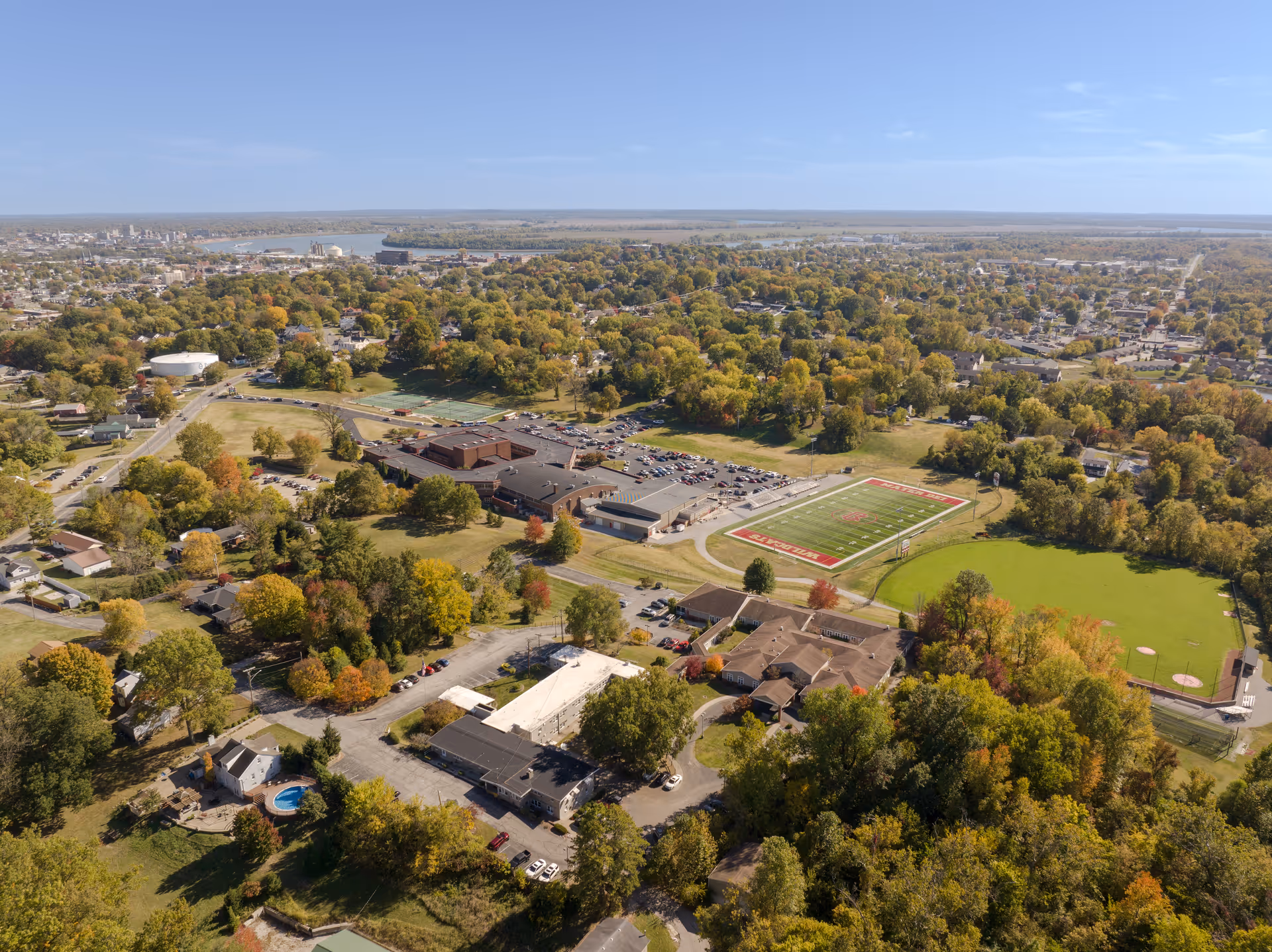 Aerial view of a suburban area with a mix of residential houses, a large school building with a football field, and surrounding trees showing fall colors under a clear blue sky.