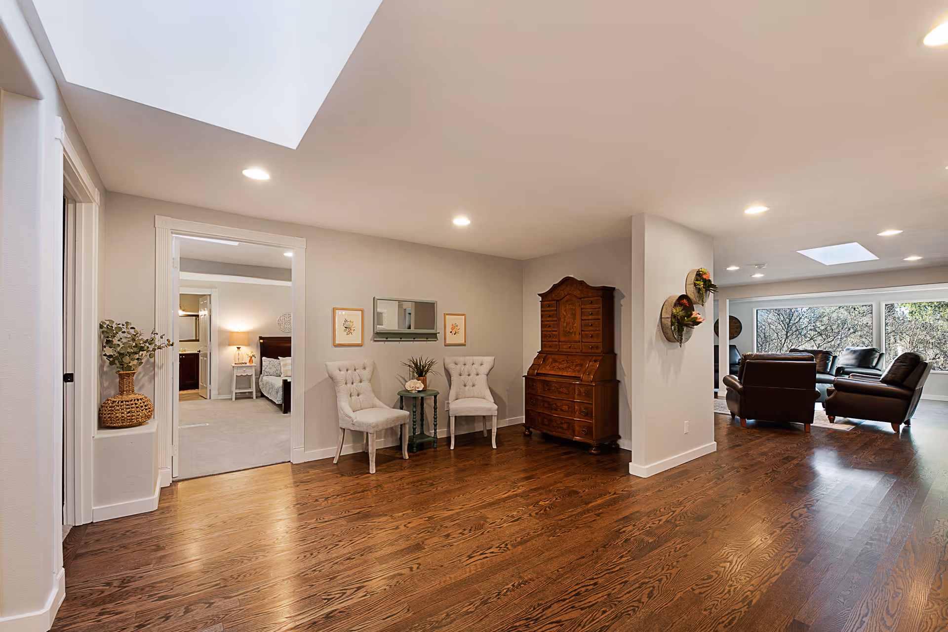 Spacious interior of a senior living facility with polished wooden floors, two upholstered chairs with a small table between them, a wooden cabinet, and a seating area with leather chairs near large windows showing outdoor greenery. A bedroom is visible through an open doorway on the left.