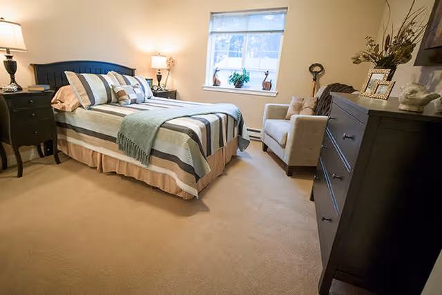 A cozy bedroom with a neatly made bed featuring striped bedding and multiple pillows. There are two bedside tables with lamps on either side of the bed. A window with blinds lets in natural light, and a comfortable armchair is placed near the window. A dark wooden dresser with decorative items and framed photos is positioned against the wall.