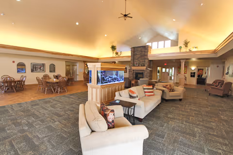 Spacious assisted living common room with sofas, a central aquarium, fireplace, and dining tables under a vaulted ceiling.