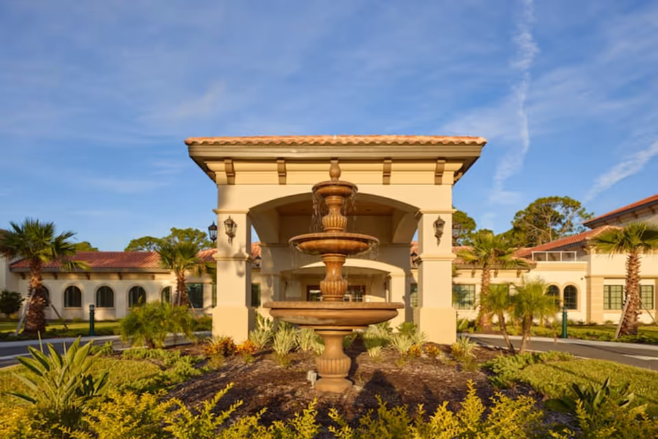Entrance of a Mediterranean-style senior living building showing a three-tier fountain in front of a covered porte-cochère and landscaped grounds.