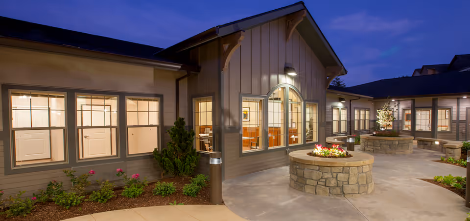 Evening view of an outdoor courtyard at Windsong at Eola Hills featuring stone planters with flowers, well-lit windows, and a paved walkway surrounded by landscaping.