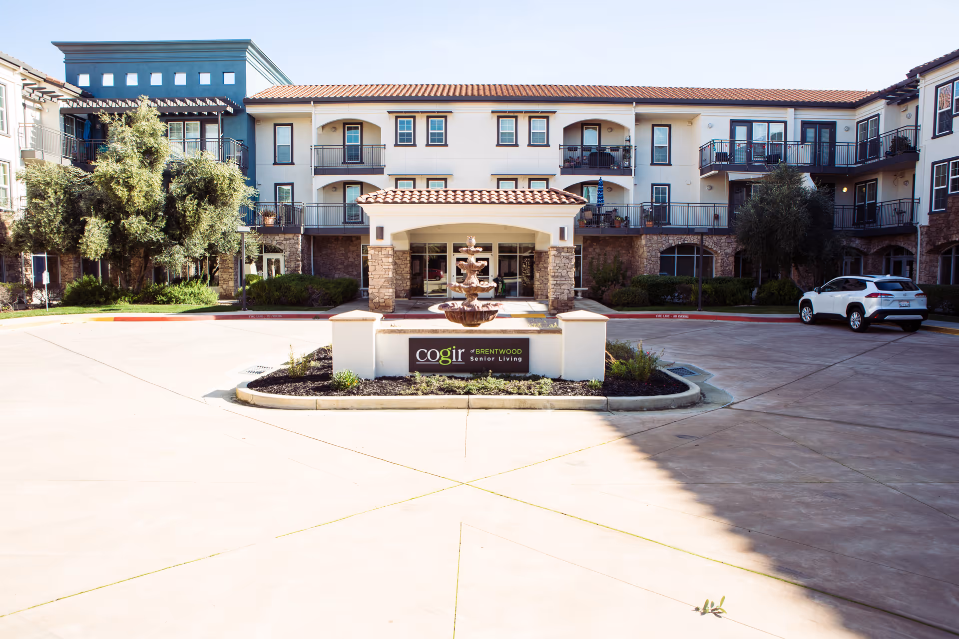 Front exterior view of Cogir of Brentwood senior living facility showing a three-story building with balconies, a central entrance with a covered portico, a decorative fountain in front, landscaped greenery, and a parked white SUV on the right side.