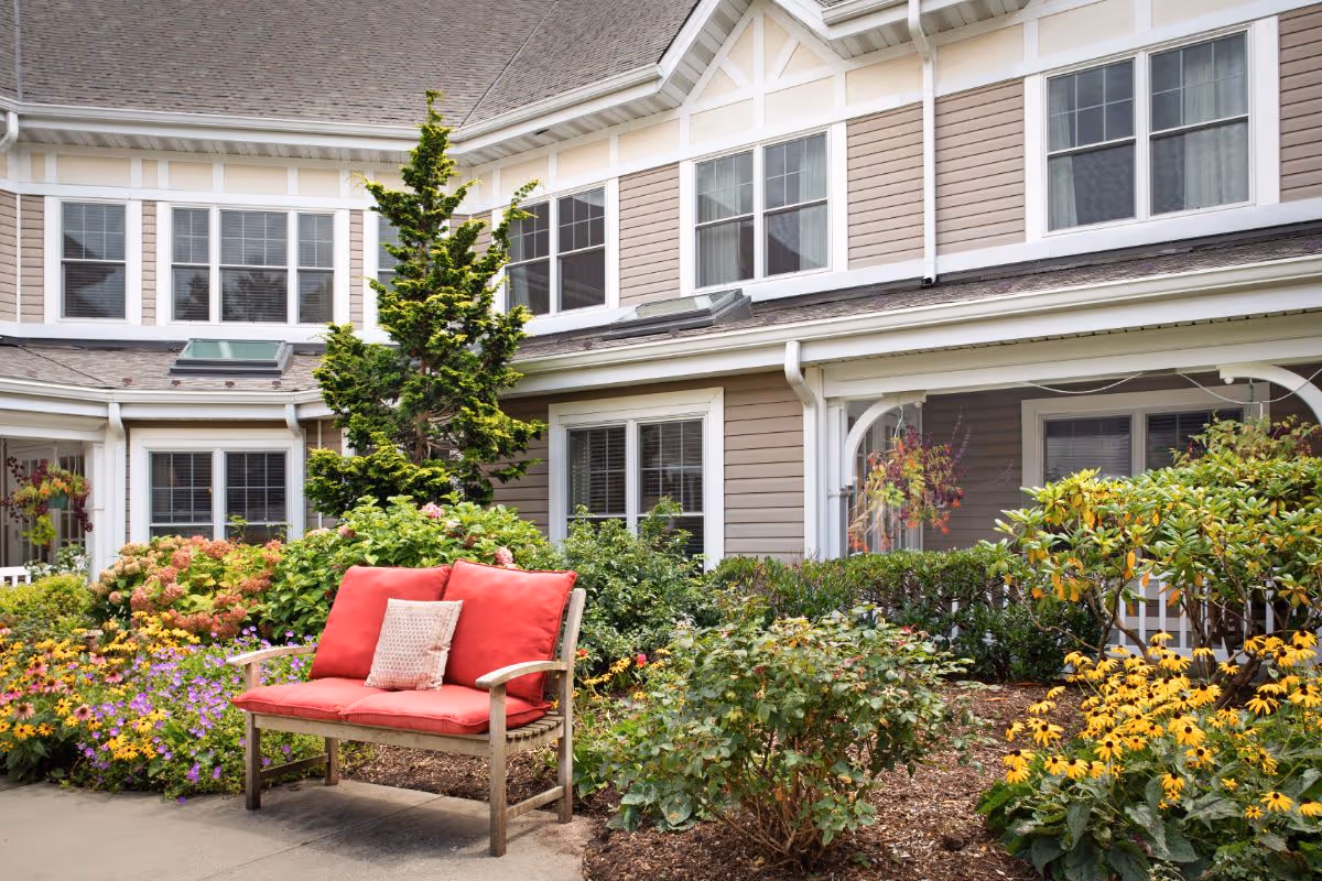 Outdoor garden area at Sunrise of North Lynbrook featuring a wooden bench with red cushions surrounded by colorful flowers and shrubs, with the building's beige exterior and multiple windows in the background.