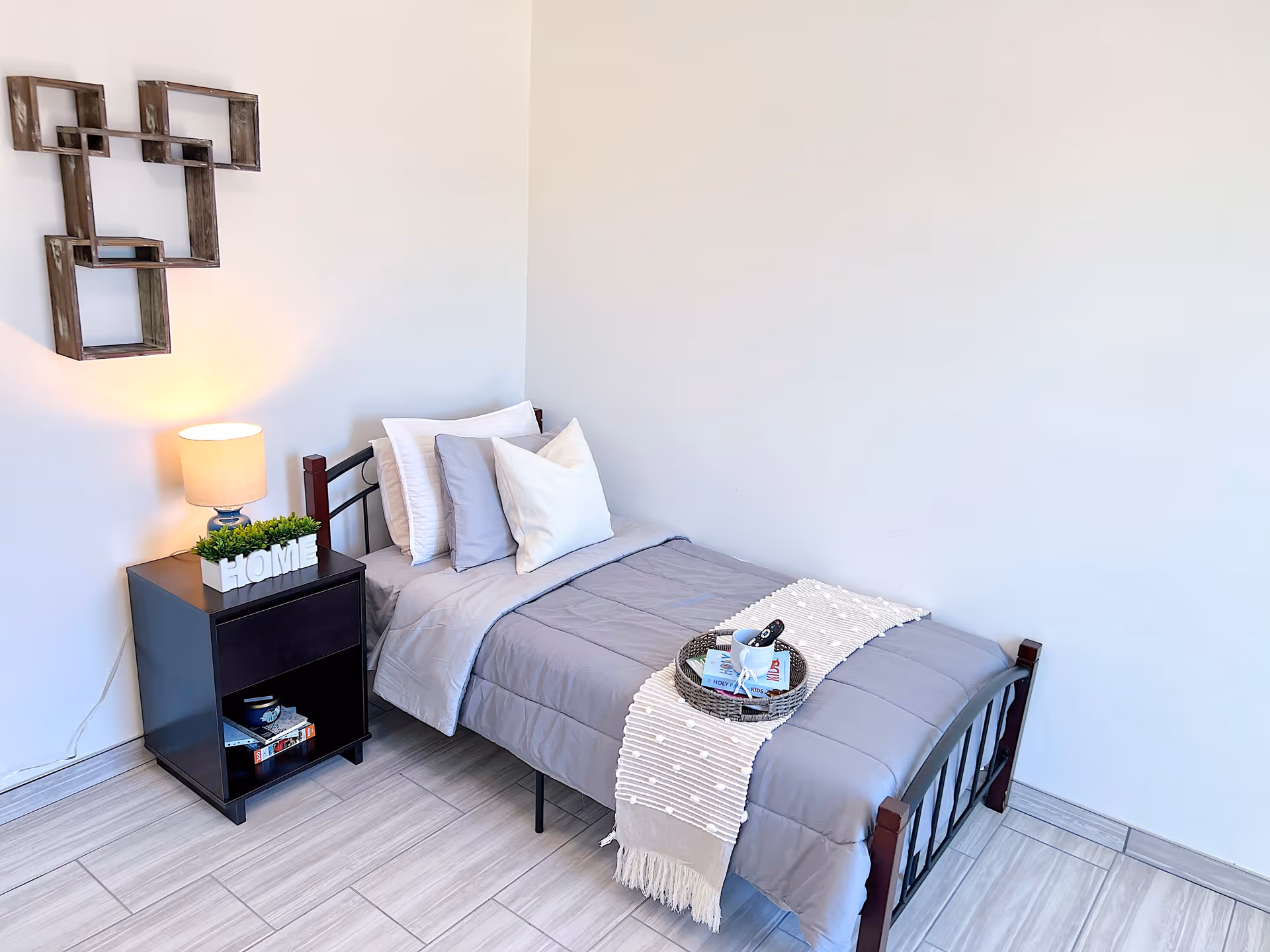 A small bedroom with a single bed made with gray bedding and multiple pillows. A decorative tray with a cup and remote controls is placed on the bed. Next to the bed is a black nightstand with a lamp, a small plant, and the word 'HOME' displayed. The wall has a geometric wooden shelf decoration, and the floor is tiled in light gray.