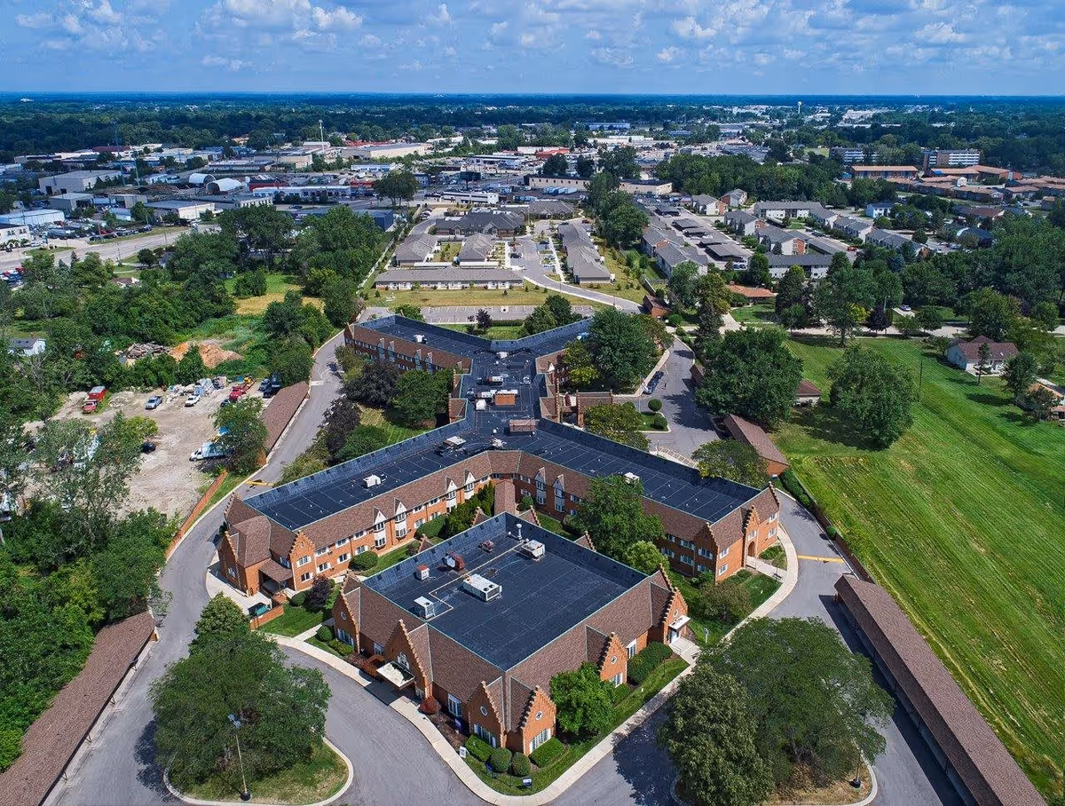 Aerial view of a senior living facility named American House East I, showing multiple connected brick buildings with black roofs, surrounded by trees, roads, parking areas, and green open spaces. The background includes a suburban area with more buildings and greenery under a partly cloudy sky.