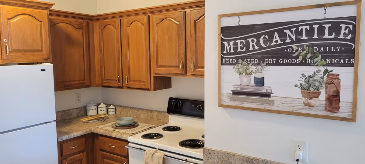 A kitchen corner with wooden cabinets, a white refrigerator, and a white electric stove. On the countertop, there are three labeled canisters for coffee, tea, and sugar, along with some plates and utensils. A framed wall art with the word 'MERCANTILE' and images of plants and jars hangs on the adjacent wall above an electrical outlet with a plugged-in charger.