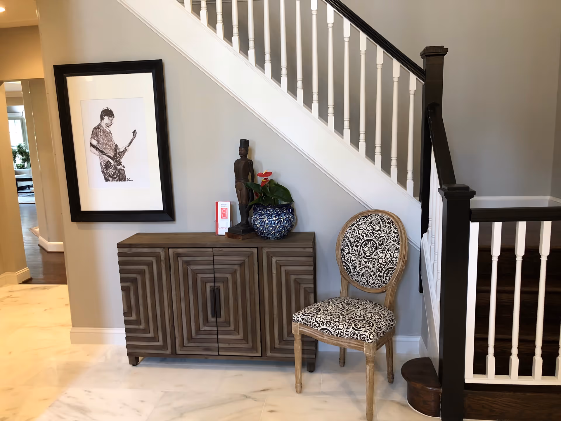 Interior view of a hallway corner with a wooden cabinet featuring a geometric pattern, a decorative statue, a blue and white flower pot with a red flower, and a patterned chair. A staircase with white spindles and dark handrails is visible on the right, and a framed black and white drawing of a person playing a guitar hangs on the wall above the cabinet.