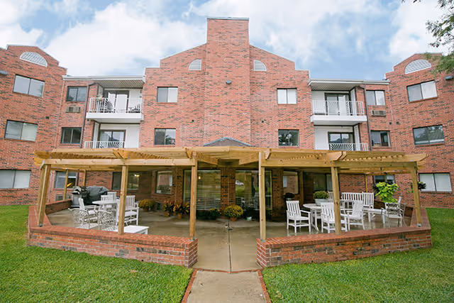 Front of a multi-story red-brick senior living building with a covered outdoor patio area furnished with white tables and chairs.