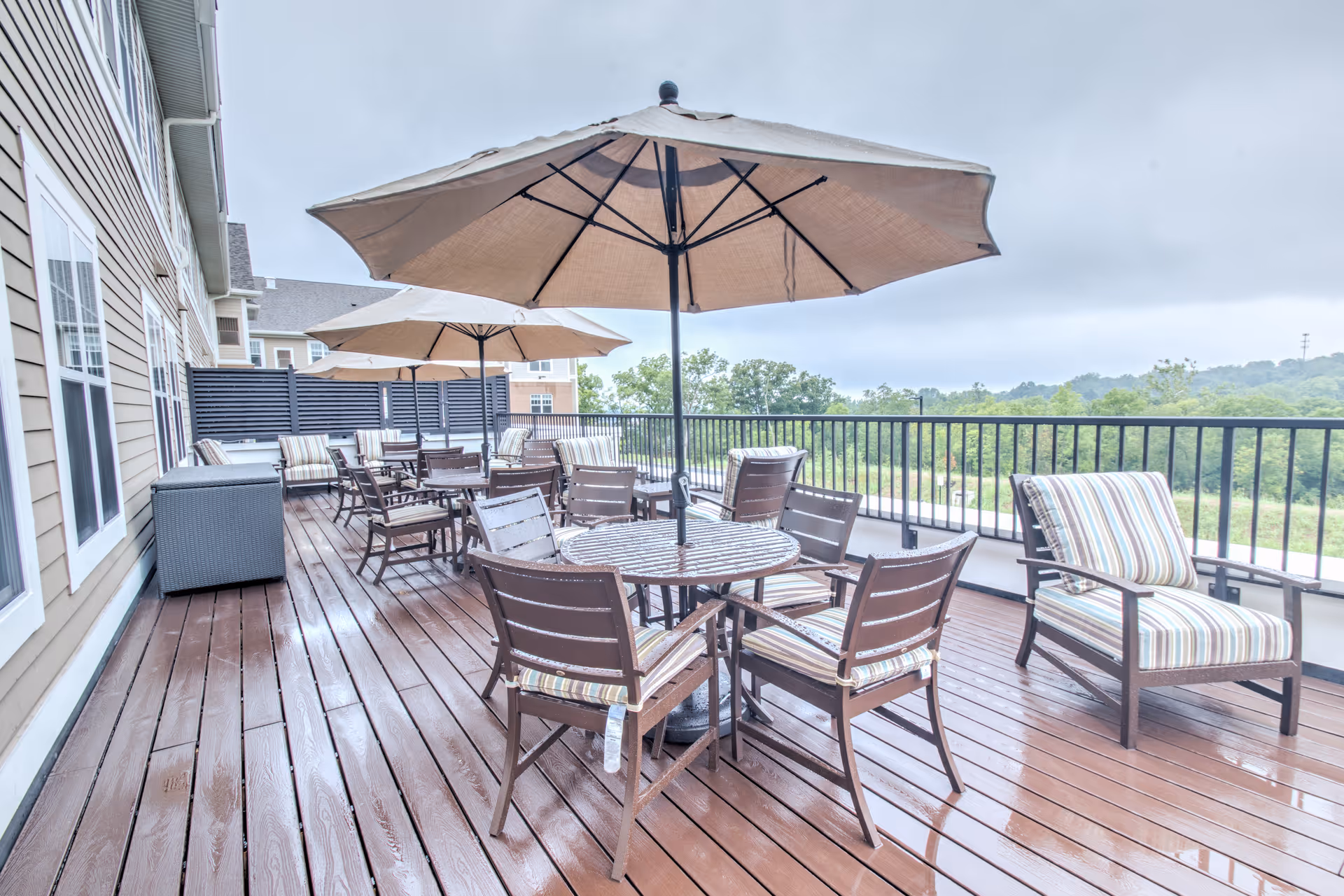 Outdoor patio area with wooden deck flooring, several round tables with chairs, and large beige umbrellas providing shade. The patio overlooks a green landscape with trees and hills in the distance under a cloudy sky.