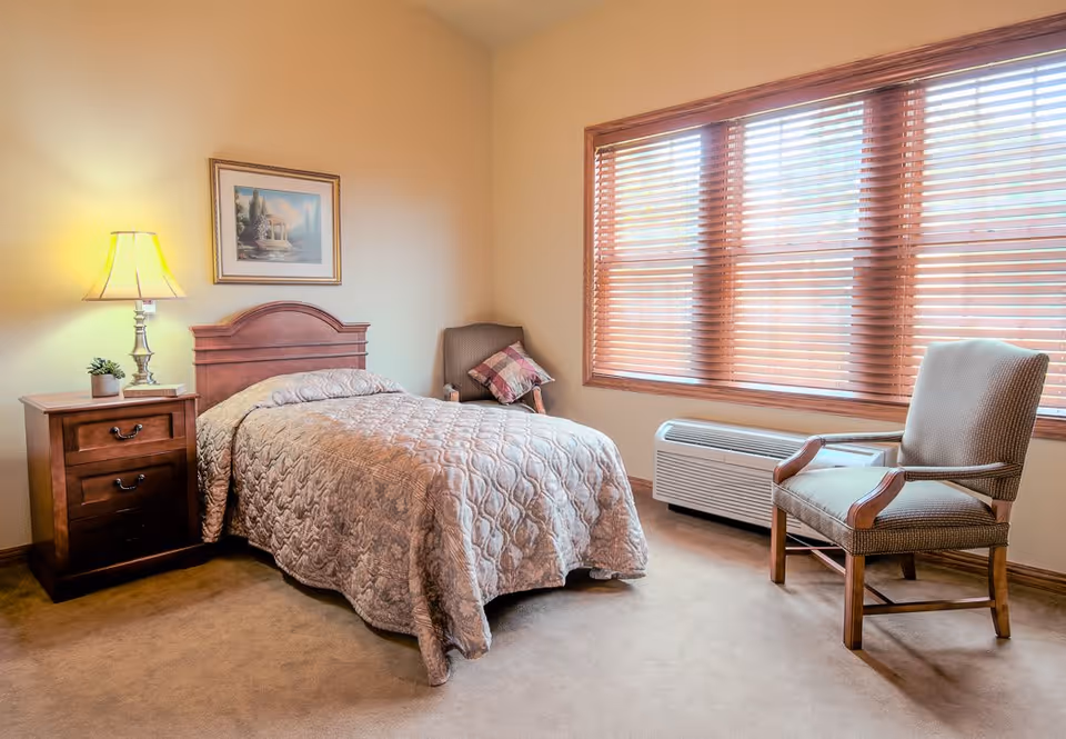 A cozy bedroom with a single bed covered in a patterned quilt, a wooden nightstand with a lamp and a small plant, a framed picture on the wall, a cushioned armchair near a large window with wooden blinds, and a heating/cooling unit beneath the window.
