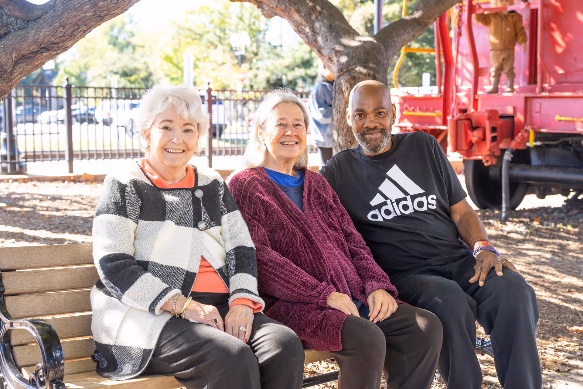 Three elderly individuals sitting on a wooden bench outdoors under a tree, smiling at the camera. Behind them is a red train car and a black metal fence with trees and greenery in the background.