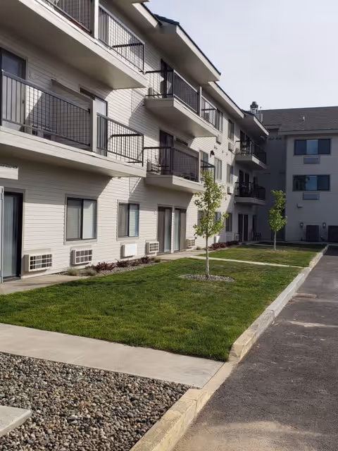 Exterior view of a multi-story apartment building with balconies facing a grassy courtyard and walkway.