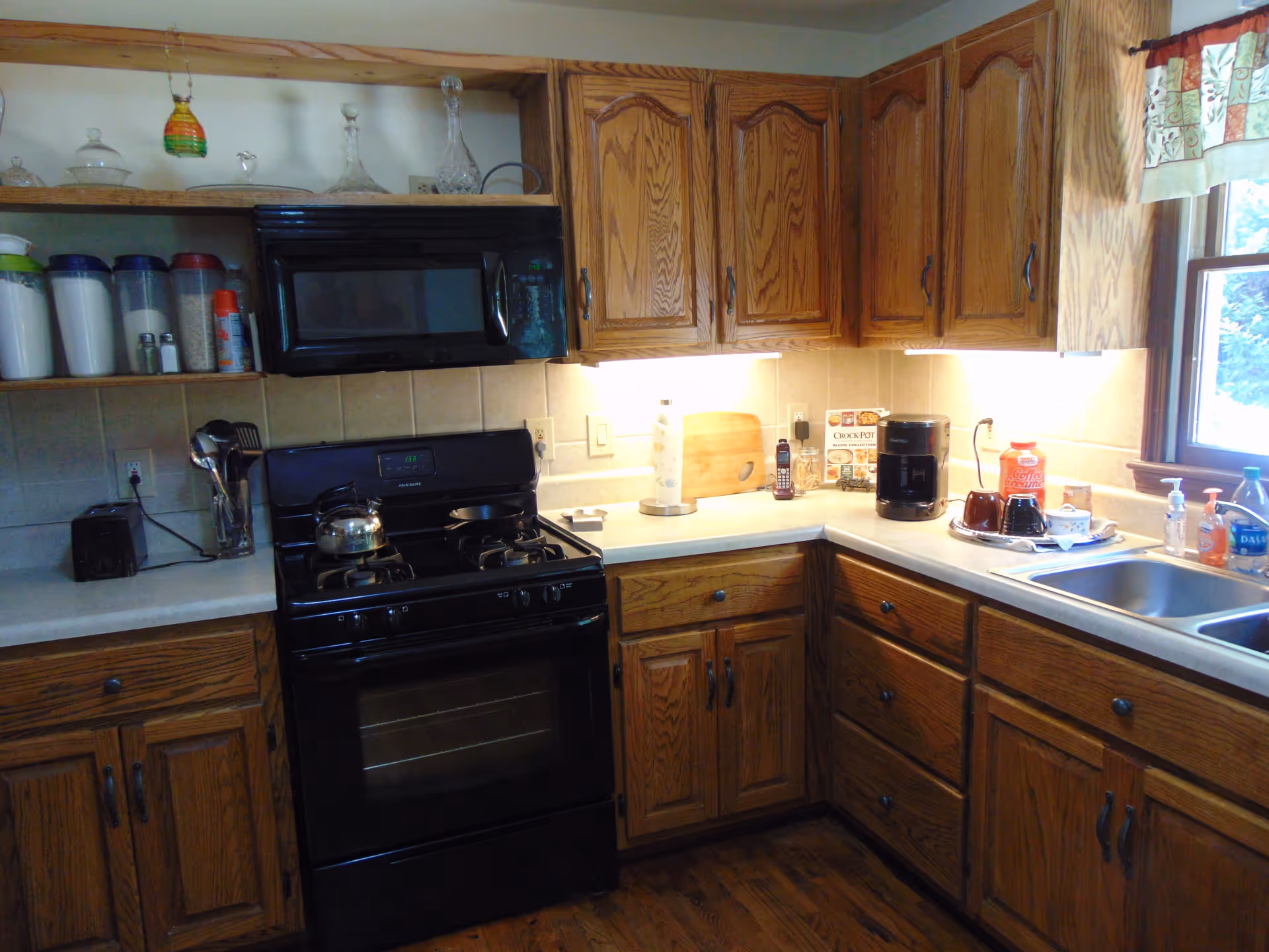 A kitchen with wooden cabinets and drawers, a black gas stove with a kettle and pan on top, a black microwave above the stove, and a countertop with various items including a coffee maker, cups, a cutting board, paper towels, and soap dispensers next to a double sink under a window with a patterned valance.