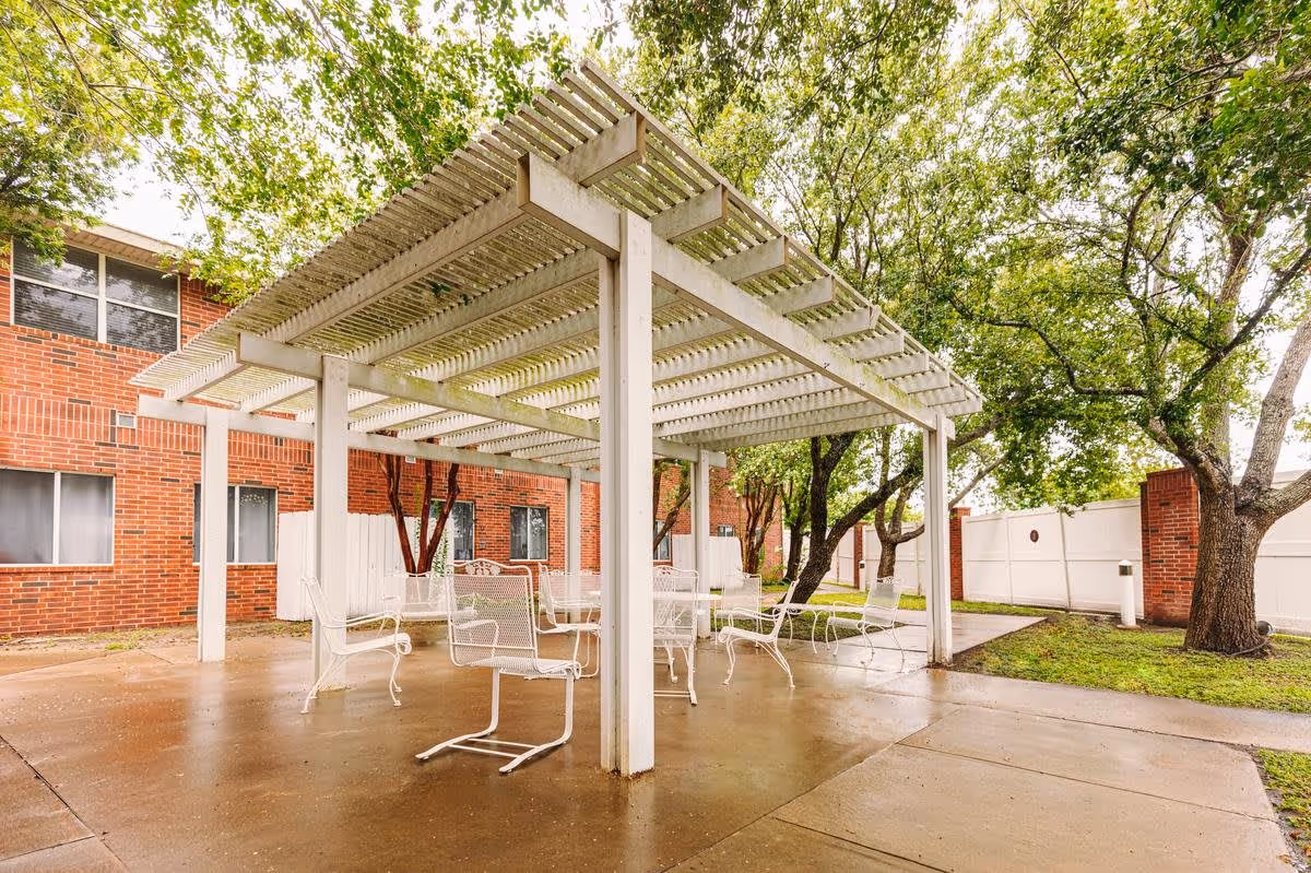 Outdoor patio area with a white pergola structure providing partial shade over several white metal chairs and tables. The patio is surrounded by green trees and grass, with a red brick building and white fencing in the background.