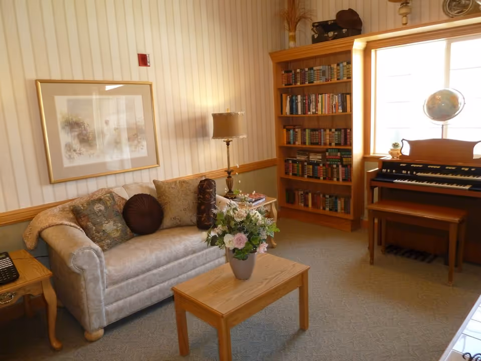 A cozy living room with a beige patterned sofa adorned with decorative pillows, a wooden coffee table with a floral arrangement, a side table with a lamp, a tall wooden bookshelf filled with books, and an organ with a bench near a large window letting in natural light.