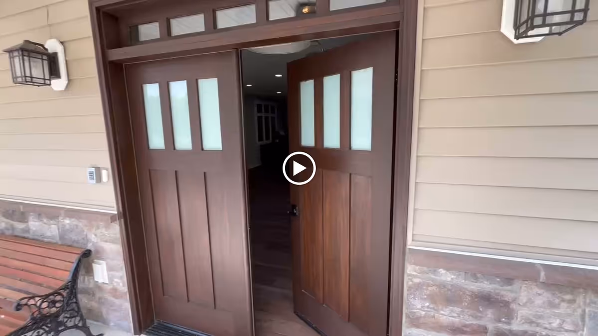 Wooden double front doors are partly open into a building entry with a bench and wall lanterns on the exterior.