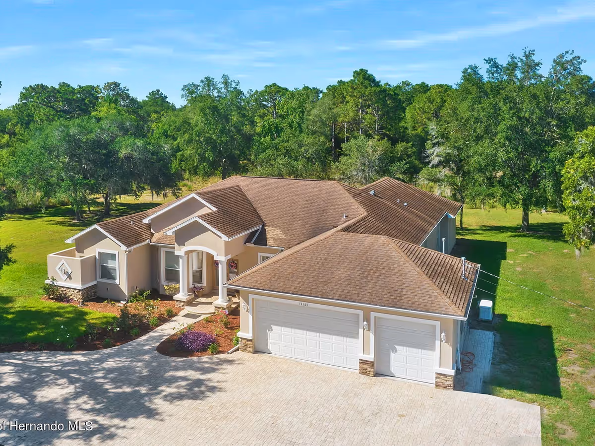 Aerial view of a single-story residential building with a brown shingle roof, beige exterior walls, and a three-car garage. The house is surrounded by green grass, landscaped flower beds, and trees in the background under a clear blue sky.