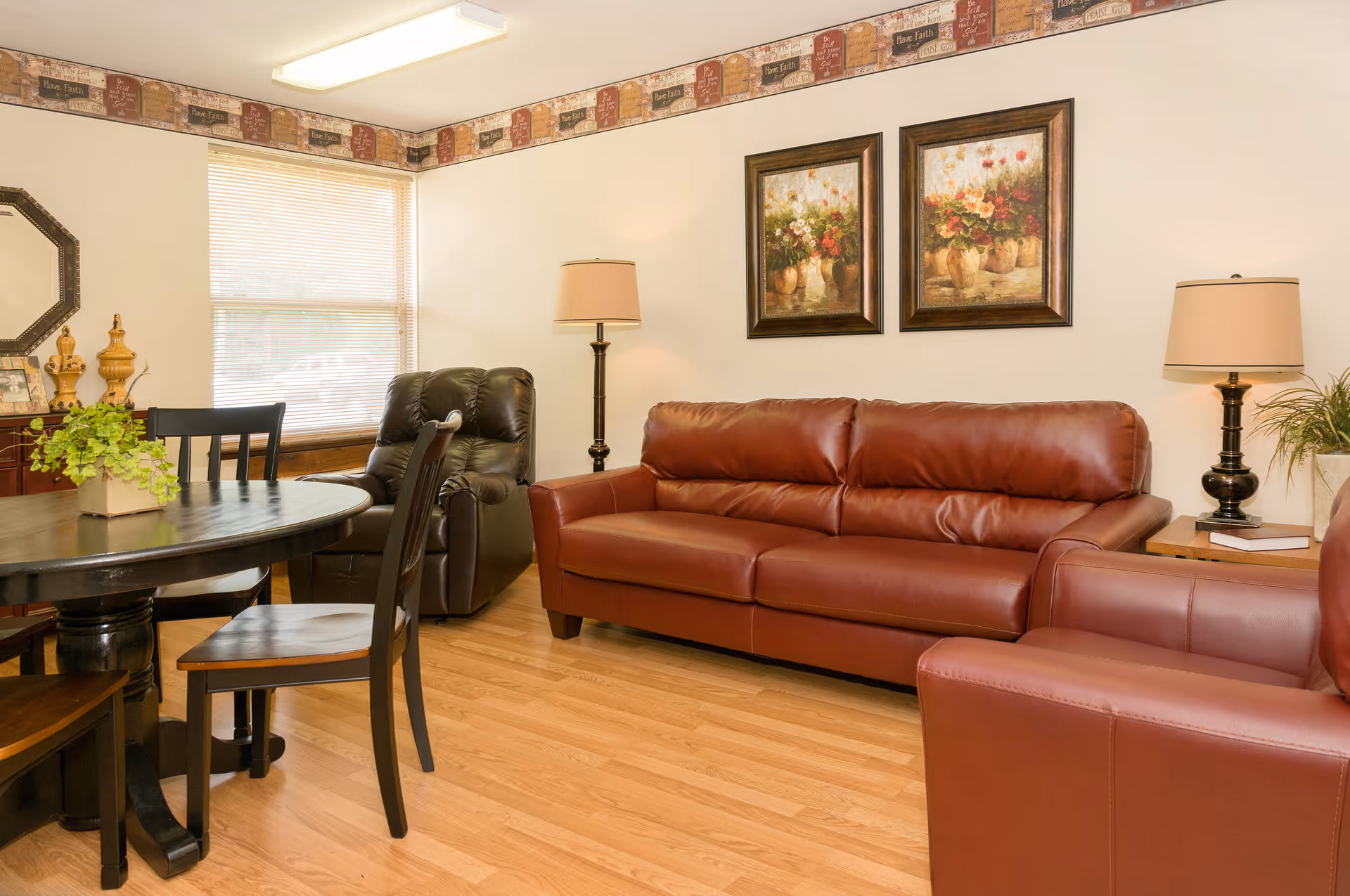 A cozy living room with a brown leather sofa and matching armchair, a black recliner chair, a round wooden dining table with chairs, two framed floral paintings on the wall, two table lamps, a window with blinds, and a decorative border near the ceiling.