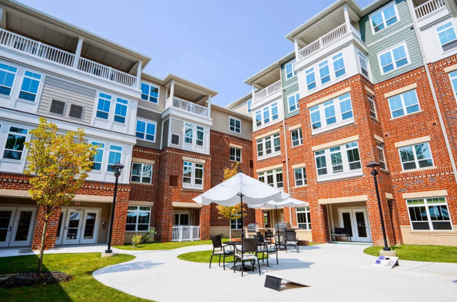 Outdoor courtyard area of a senior living facility with brick and siding multi-story buildings surrounding a concrete patio. The patio has tables with umbrellas and chairs, small trees, and a cornhole game set up on the ground. The sky is clear and blue.