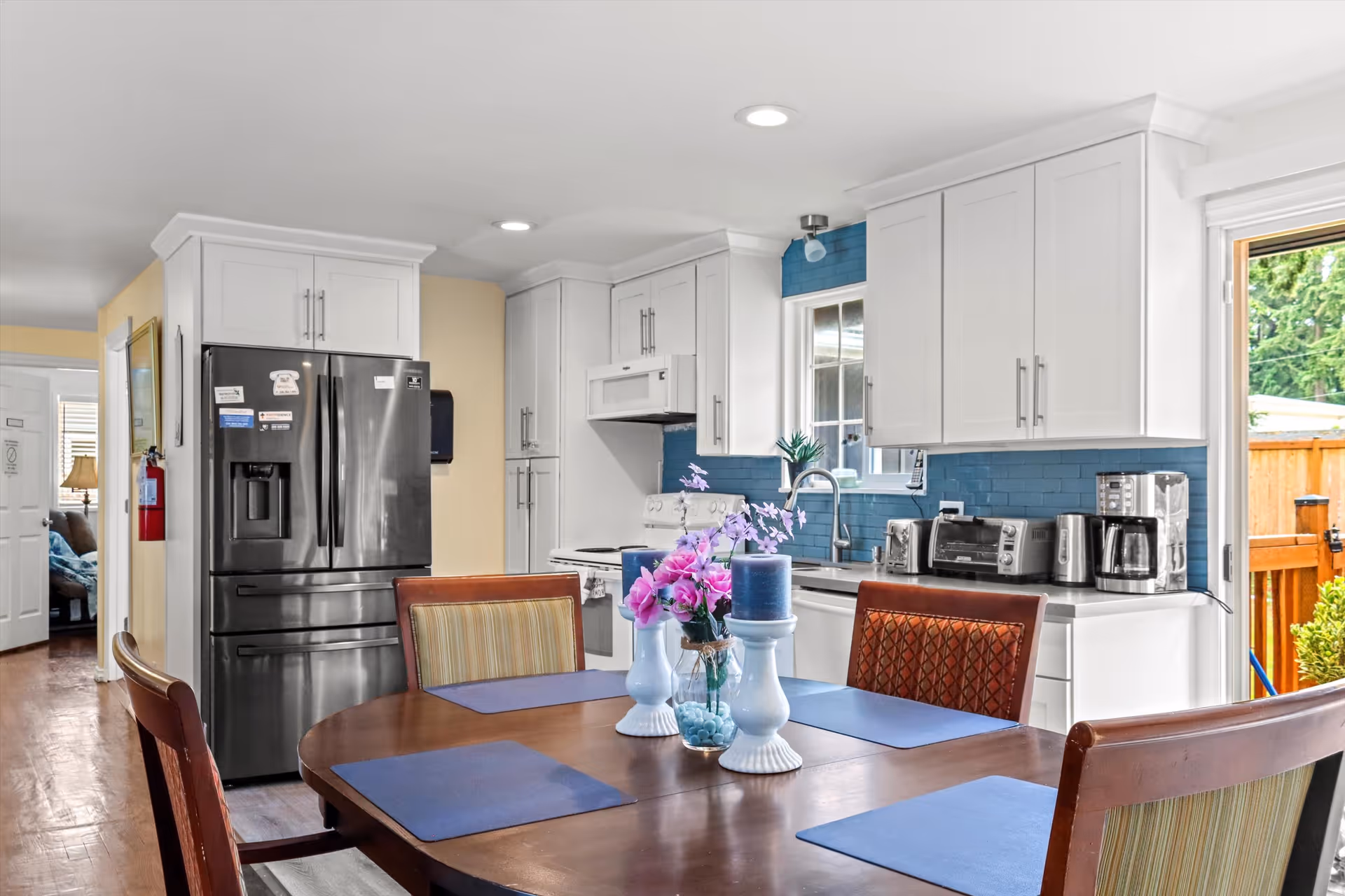 A bright kitchen and dining area featuring white cabinets, stainless steel refrigerator, stove, microwave, and various small appliances on the countertop. A wooden dining table with four chairs is set with blue placemats and decorative items including flowers and candles. A window above the sink and a door leading outside provide natural light.