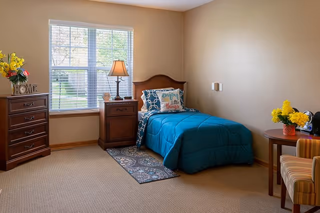 Sunlit bedroom with a single bed in teal bedding, wooden nightstand and dresser, window with blinds, and a small table with a striped chair.