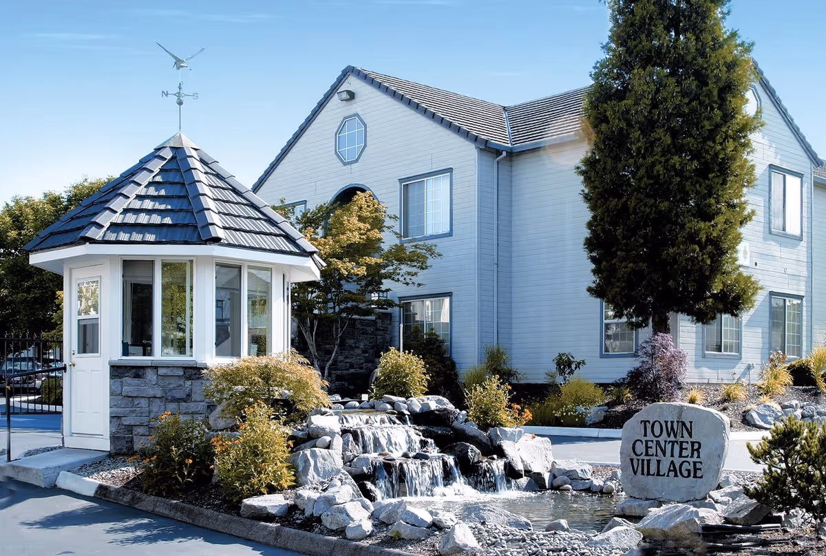 Exterior view of a senior living facility named Town Center Village featuring a small stone and white guardhouse with a weather vane on top, a cascading rock waterfall, landscaped bushes, and a large tree in front of a two-story building with multiple windows.