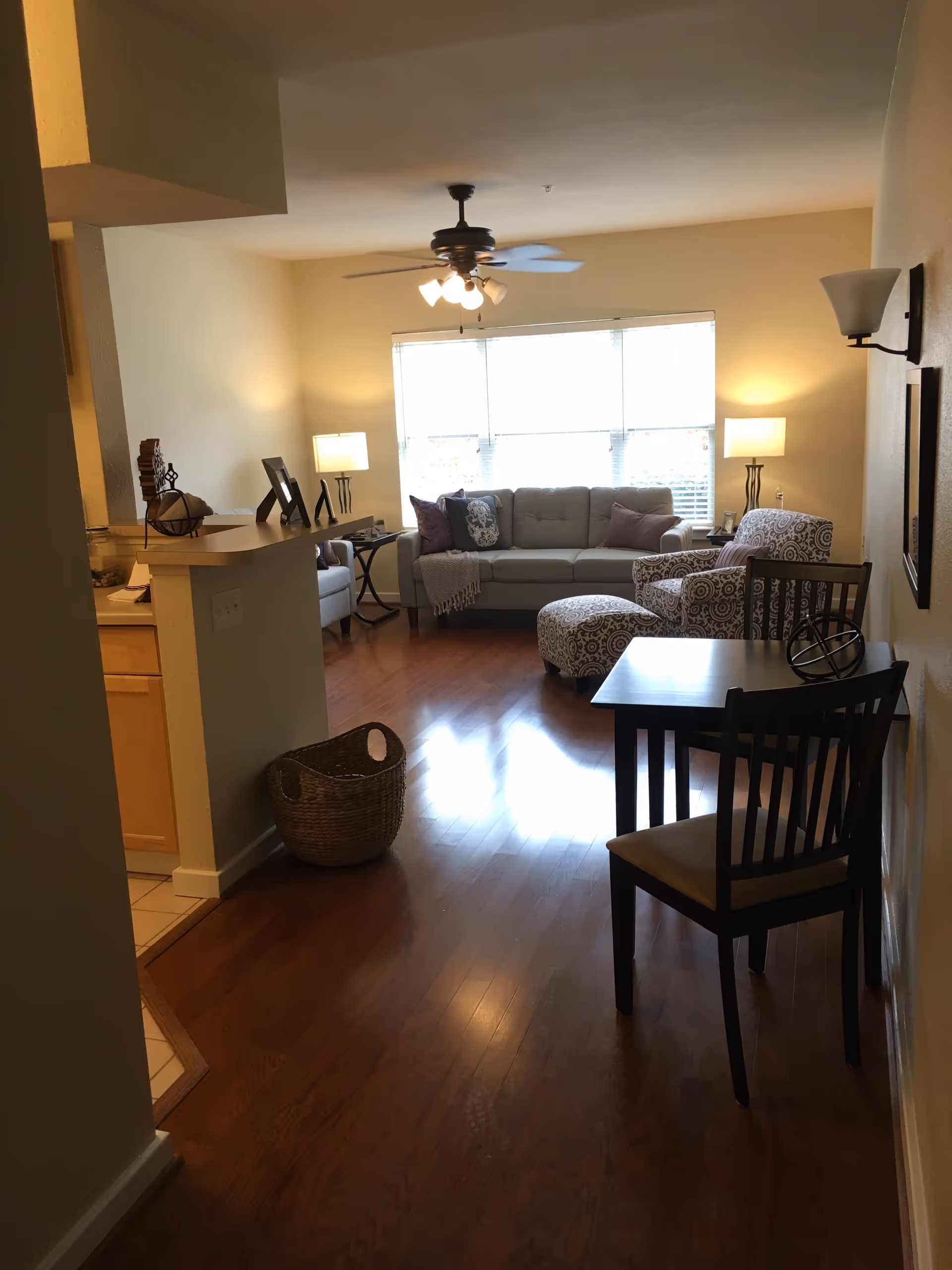 A cozy living room with wooden flooring, a ceiling fan with lights, a large window with blinds, a gray sofa with pillows, a patterned armchair with matching ottoman, two side tables with lamps, a small dining table with two chairs, and a woven basket on the floor near a partial kitchen counter.