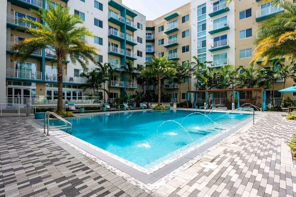 Outdoor swimming pool in the courtyard of a multi-story residential building with palm trees, lounge chairs, and water fountains in the pool.