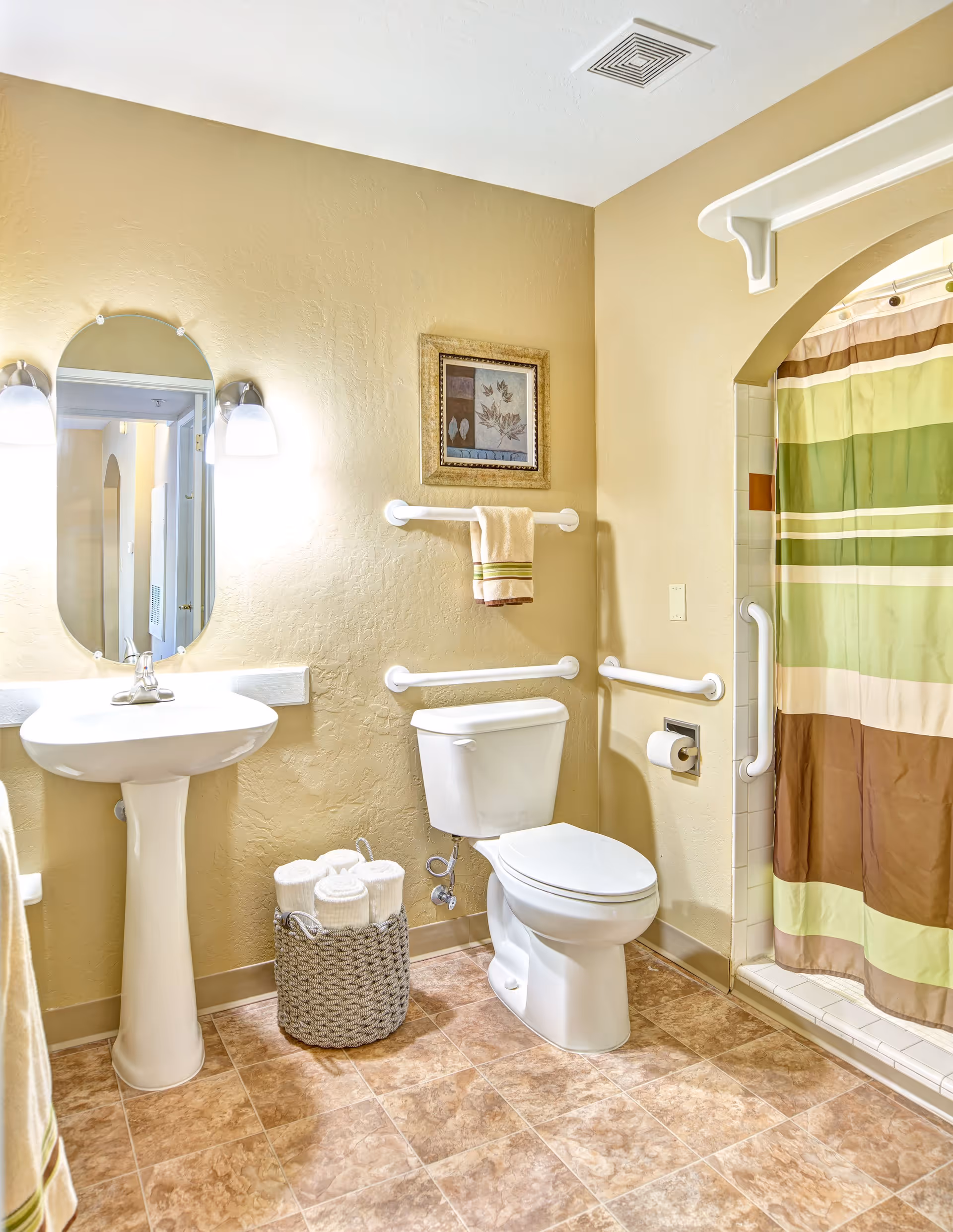 A clean and accessible bathroom with beige walls and tiled floor. It features a white pedestal sink with an oval mirror above it, two wall-mounted lights on either side of the mirror, a white toilet with grab bars around it, and a shower with a striped green, brown, and beige curtain. A basket with rolled white towels is placed on the floor next to the toilet, and a framed picture hangs above the towel rack.