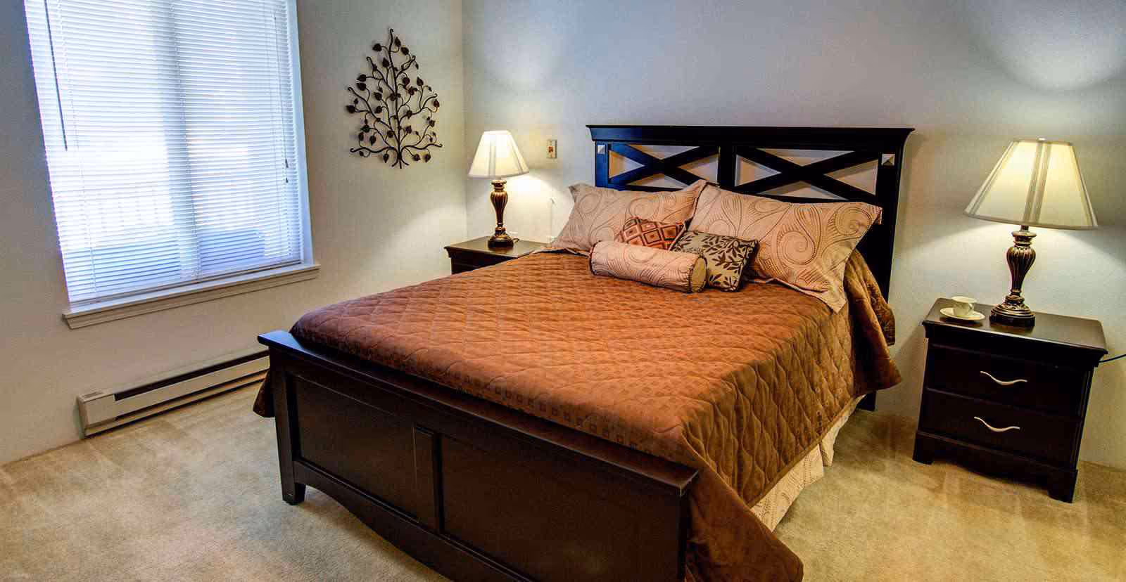 A bedroom with a dark wooden bed frame and a brown quilted bedspread. The bed is decorated with multiple pillows in various patterns. There are two matching dark wooden nightstands on either side of the bed, each with a lamp. A window with blinds is on the left wall, and a decorative metal wall art piece is mounted above the left nightstand.