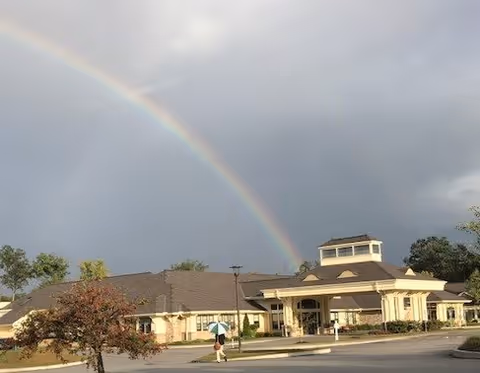 A single-story building with a large entrance and a cupola on the roof under a cloudy sky. A rainbow arcs across the sky above the building. There is a person walking on the sidewalk in front of the building, holding an umbrella. Trees and a parking lot are visible around the building.