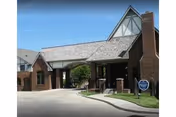 Entrance to a senior living facility with a covered driveway, brick pillars, and a peaked roof structure. The building has a mix of brick and light-colored siding with a chimney visible. There is a blue sign on the lawn near the driveway.