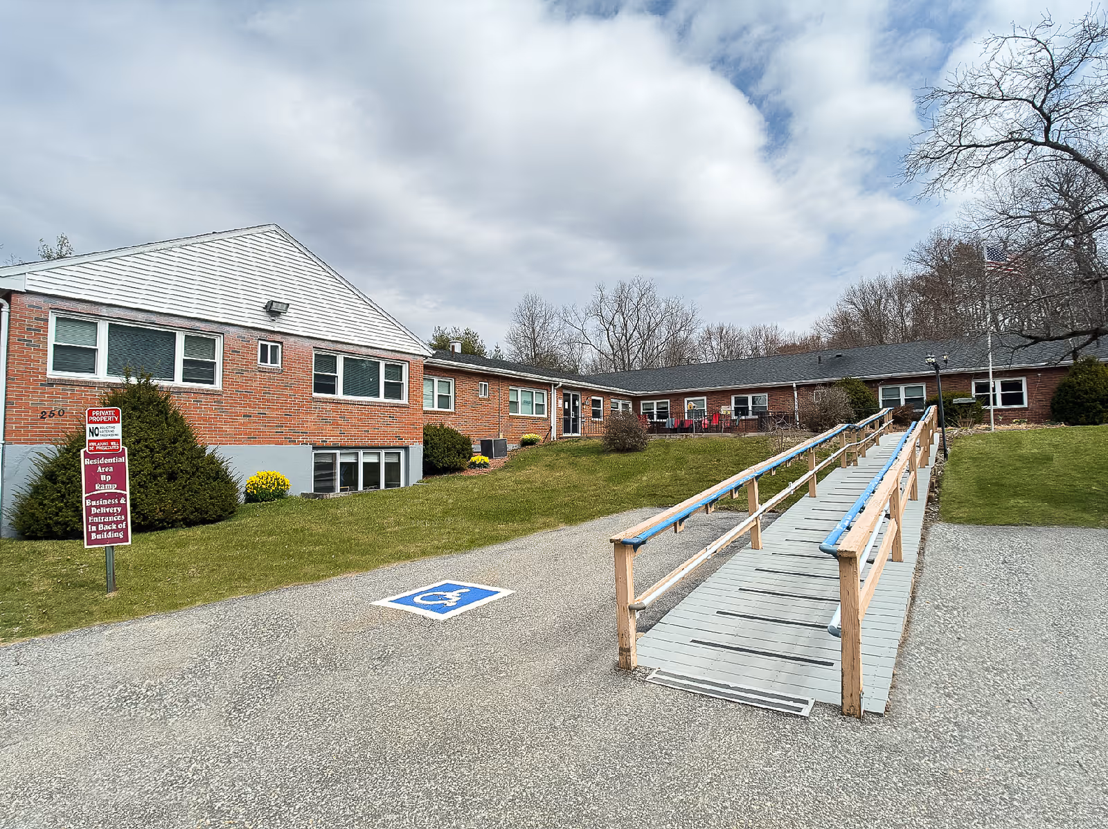 Exterior view of Palmer Healthcare Center showing a single-story brick building with multiple windows, a wooden wheelchair ramp leading to the entrance, a handicapped parking space, and a grassy area with bushes and trees under a partly cloudy sky.