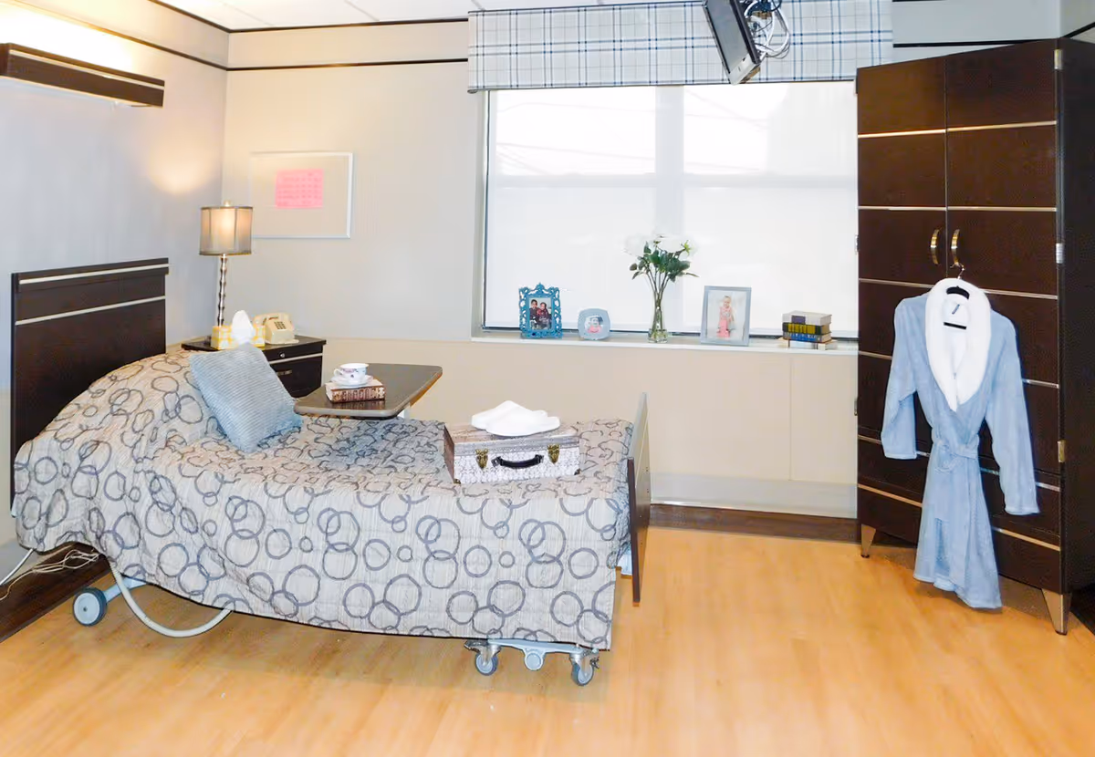 A well-lit bedroom in a healthcare facility featuring a hospital-style bed with patterned bedding and a gray pillow. A small table on wheels is placed over the bed holding a cup and a book. Next to the bed is a nightstand with a lamp and a telephone. A large window with a blind lets in natural light, and the windowsill holds framed photos, a vase with white flowers, and some books. A dark wooden wardrobe stands in the corner with a light blue robe hanging on its handle. The room has light wooden flooring and neutral-colored walls.