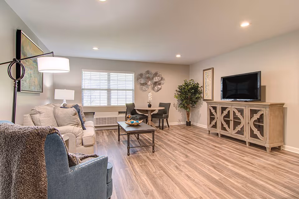 A bright and spacious living room with light wood flooring, a beige sofa with cushions, a blue armchair with a throw blanket, a wooden coffee table, a round dining table with two chairs, a large window with blinds, a decorative wall art, a potted plant, and a wooden TV stand with a flat-screen television.