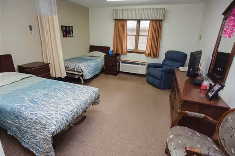 A senior living facility bedroom with two single beds covered in blue patterned bedspreads. The room has beige walls and carpet, a window with beige curtains, a blue recliner chair, a wooden dresser with a TV and framed photos, and a wooden chair with a floral cushion.