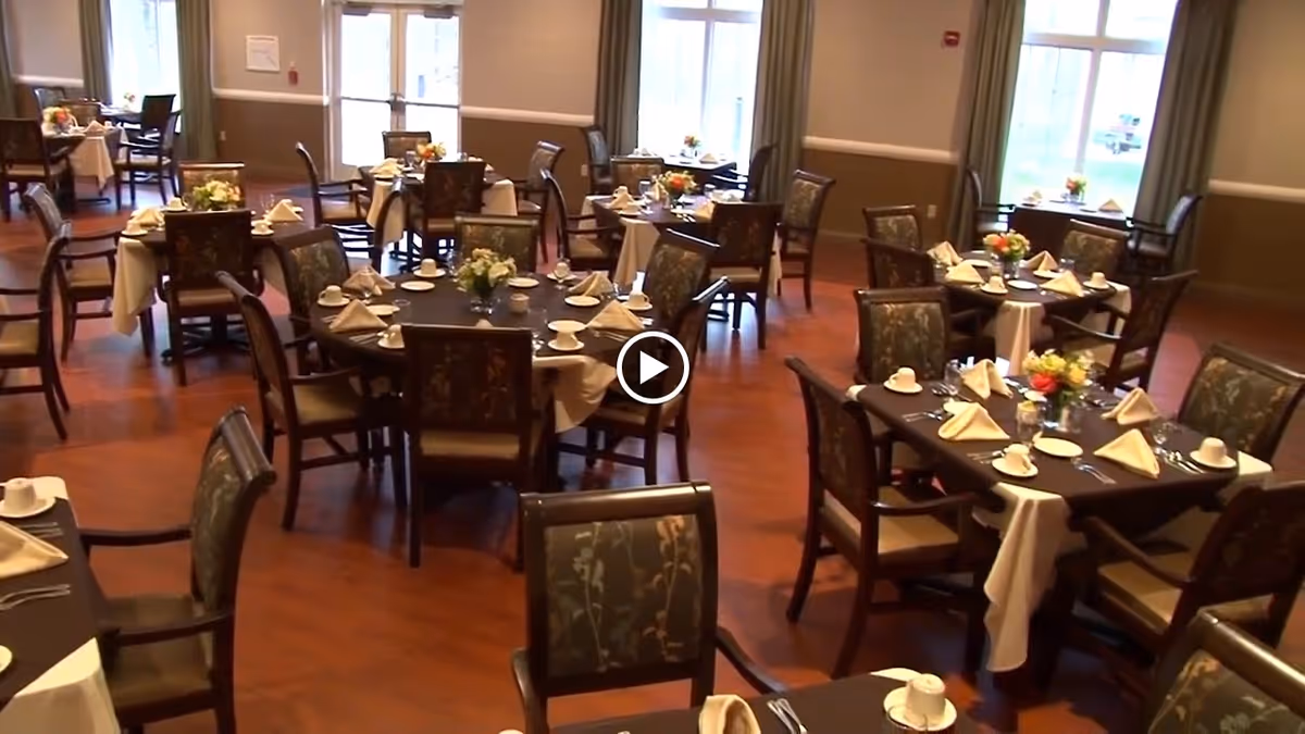 A dining room in a senior living facility with multiple round and square tables set with white napkins, cups, plates, and floral centerpieces. The room has wooden flooring, large windows with curtains, and chairs with patterned upholstery.