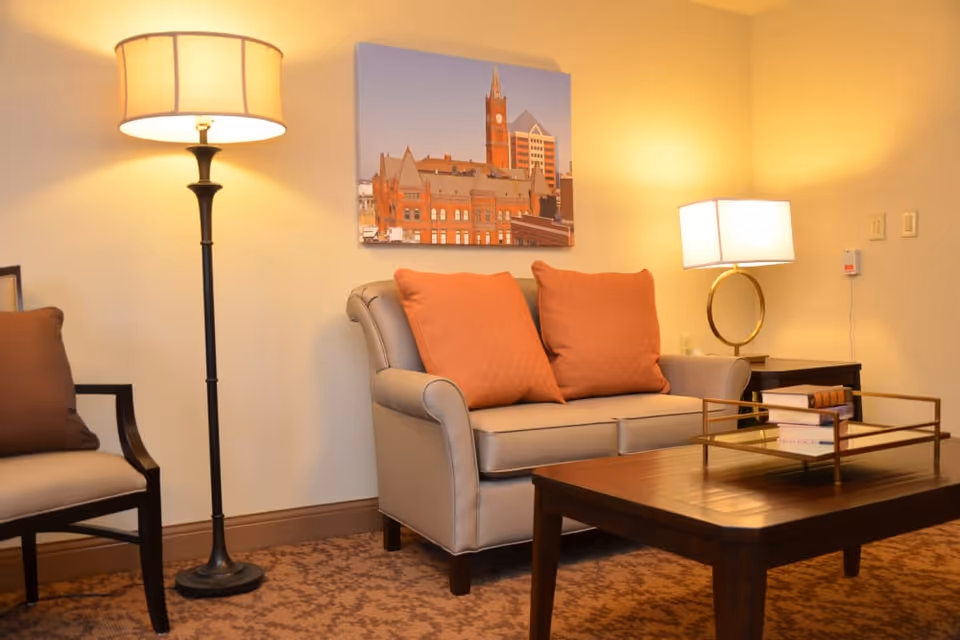 A cozy living room area with a beige loveseat featuring two orange cushions, a wooden coffee table with books on a tray, a floor lamp with a beige shade, a side table with a lamp, and a framed picture of a cityscape on the wall.
